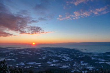 A sunrise seen from snow-capped peak of Schoeckl in Austrian Alps. The sky is bursting with orange and pink. Distant view on Graz, shrouded with fog. Winter wonderland. Day break. Calmness