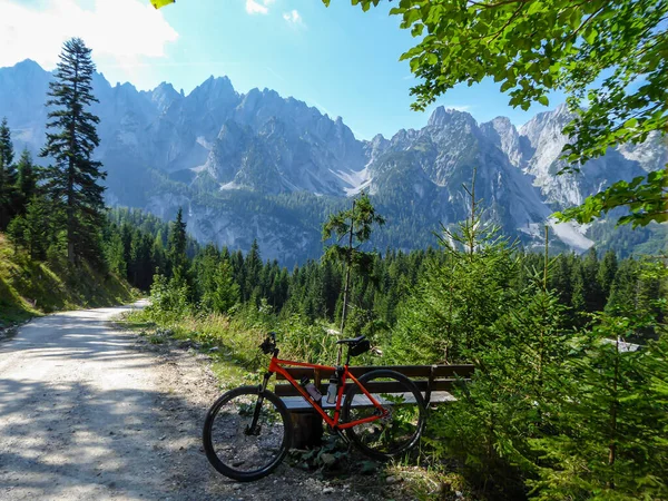 An orange mountain bike leaning against a bench next to a gravelled road in the mountains with the view on high Alps in the region of Gosau, Austria. The chains in the back are stony and barren. Calm