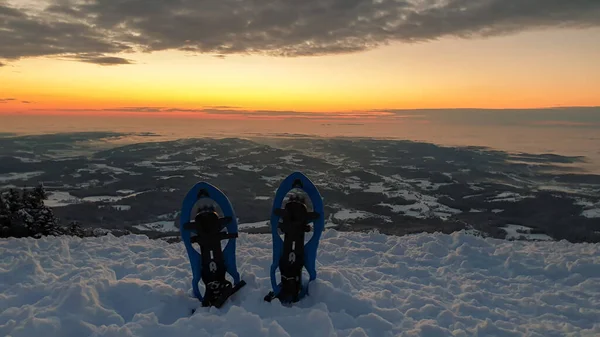 A pair of snow shoes stuck into powder snow at the peak of Schoeckl in Austrian Alps, with the view on sun rising above the horizon. The sky is bursting orange. Winter wonderland. Day break. City view