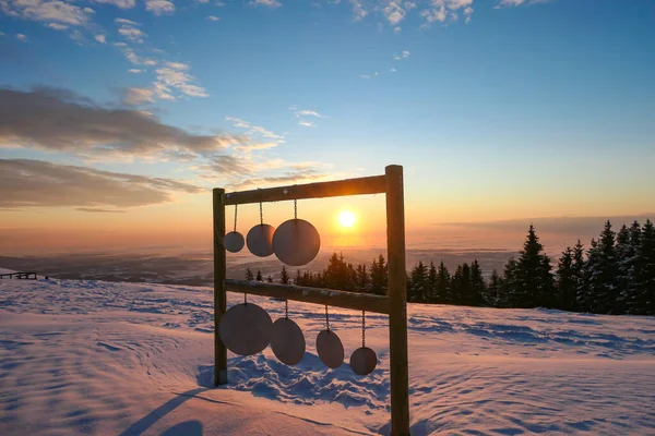 A wooden frame with round plates hanging from it located at the top of snow covered peak of Schoeckl, Austrian Alps, captured during the sunrise. The sky is bursting orange. Winter landscape. Daybreak