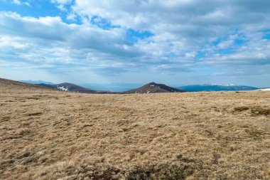 A panoramic view on an Alpine chain in Austria. The vast pasture has golden colors. There are other mountain chains in the back. A narrow pathway leading to the top. Overcast. Serenity and calmness