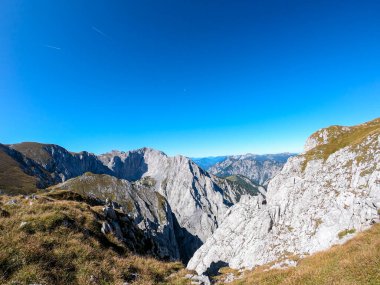 A panoramic view on Hochschwab mountain chains from the pathway leading to Hohe Weichsel. There is a vast pasture on top of a mountain, slowly turning golden. Clear view. Blue sky above. Autumn vibe