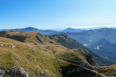 A panoramic view on Hochschwab mountain chains from the pathway leading to Hohe Weichsel. There is a vast pasture on top of a mountain, slowly turning golden. Clear view. Blue sky above. Autumn vibe