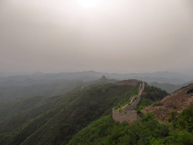 A panoramic view on an unrenewed Gubeikou part of Great Wall of China. The wall is spreading on tops of mountains. Many watchtowers on the peaks. Dense forest around it. World wonder. Tradition