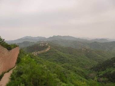 A panoramic view on an unrenewed Gubeikou part of Great Wall of China. The wall is spreading on tops of mountains. Many watchtowers on the peaks. Dense forest around it. World wonder. Tradition