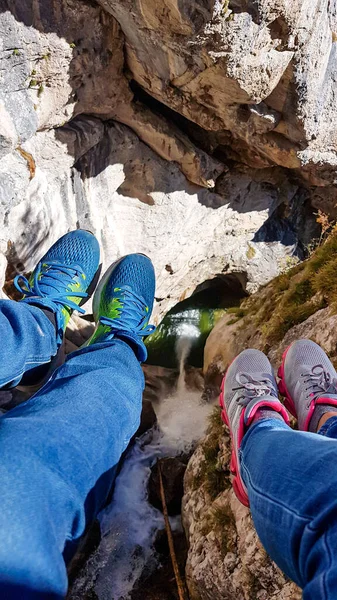 Two pairs of legs hanging from a ladder. There is a high mountain gorge below. Adventure park in Austrian Alps. There is a waterfall flowing in the gorde. Natural phenomenon