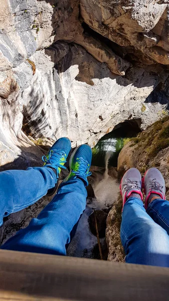 Two pairs of legs hanging from a ladder. There is a high mountain gorge below. Adventure park in Austrian Alps. There is a waterfall flowing in the gorde. Natural phenomenon