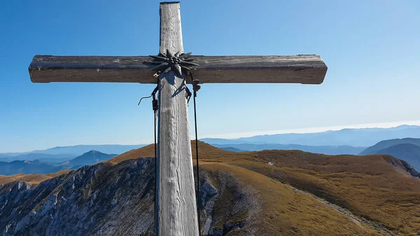A wooden cross on top of Hohe Weichsel, Alpine peak in Austria. The cross is leaning, as if it was going to fall. There are endless mountain chains behind it. Early fall. The slopes are turning golden