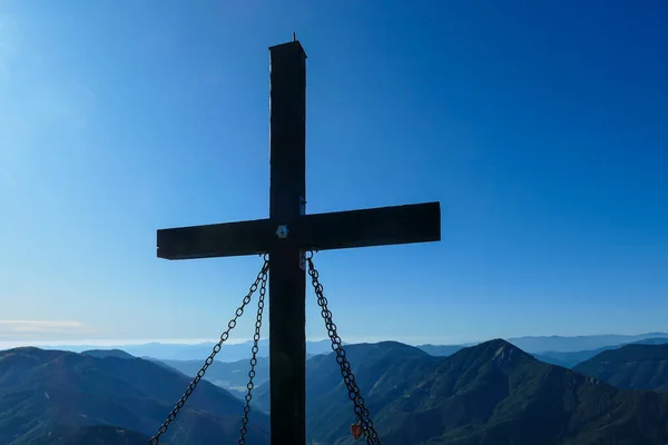 A wooden cross on top of Hohe Weichsel, Alpine peak in Austria. The cross is supported by 4 metal ropes. There are endless mountain chains behind it. Early fall. The slopes are turning golden