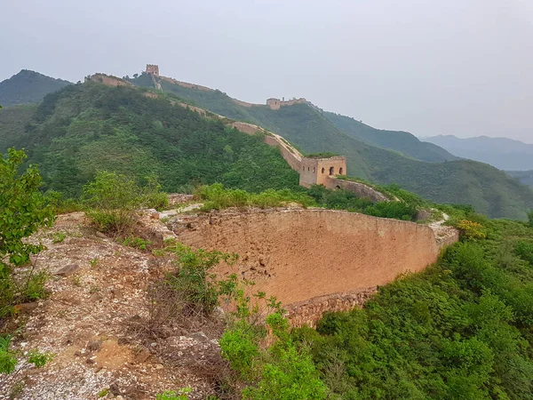 A panoramic view on an unrenewed Gubeikou part of Great Wall of China. The wall is spreading on tops of mountains. Many watchtowers on the peaks. Dense forest around it. World wonder. Tradition