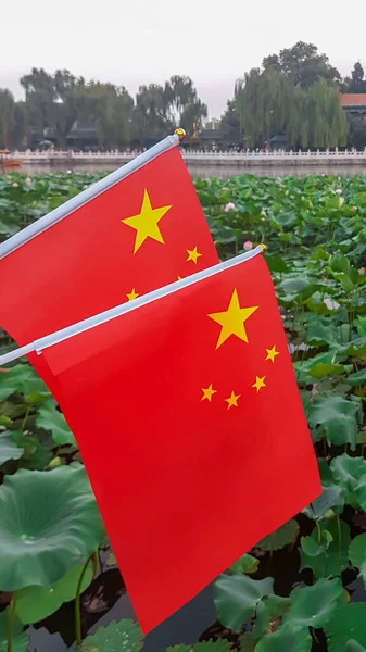 Two Chinese flags waving gently on the wind in Beihai Park in Beijing, China. Under the flags there is a small lake, overgrown with water lilies. There is a white marble railing along the lake.