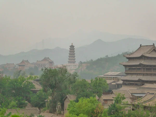 A panoramic view on a small village in northern China. A tall pagoda striking above the village.  There is a dense forest around. Rural areas. Tradition meets modernisation. Overcast and air pollution
