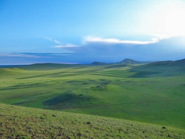 A panoramic view on a hilly landscape of Xilinhot in Inner Mongolia. Endless grassland with a few wildflowers between. The sun starts to set, coloring the sky orange. Blue sky with thick, white clouds