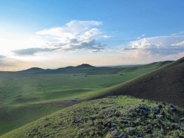 A panoramic view on a hilly landscape of Xilinhot in Inner Mongolia. Endless grassland with a few wildflowers between. The sun starts to set, coloring the sky orange. Blue sky with thick, white clouds