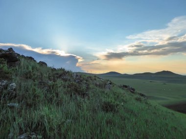 A panoramic view on a hilly landscape of Xilinhot in Inner Mongolia. Endless grassland with a few wildflowers between. The sun starts to set, coloring the sky orange. Blue sky with thick, white clouds