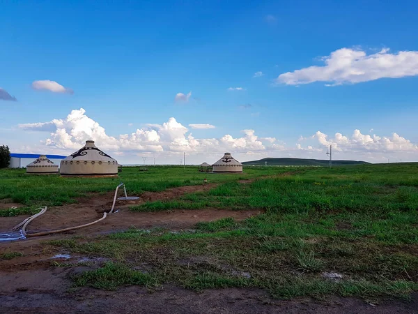 A few white, traditional yurts located on a pasture in Xilinhot in Inner Mongolia. Endless grassland. Blue sky with a few thick, white clouds. A long garden hose in front. Nomadic way of life.