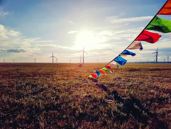Colorful prayer flags attached to a heap of stones (aobao) on a vast grassland in Xilinhot, Inner Mongolia. The sun is setting behind the horizon. Golden hour. A field of wind turbines in the back
