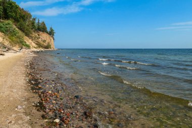 A panoramic view on the costal line in Gdynia, Poland, seen from a sandy beach. Calm Baltic Sea reflecting sunbeams from its calms surface. High trees overgrowing the tall cliffs. Idyllic