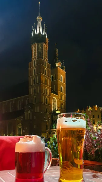 A dark and wheat beer standing on a small table on the square in Cracow, Poland, with the view on St. Mary\'s Basilica. The two bell towers od the basilica are towering above the city. Dark night