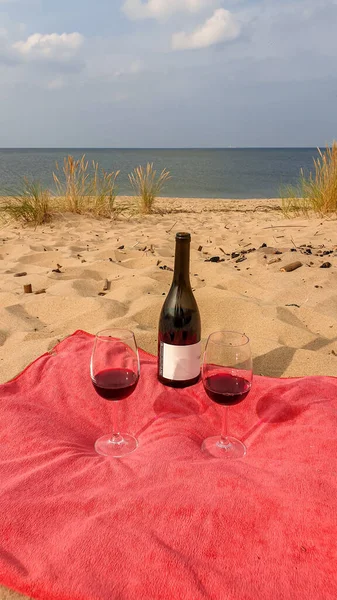 A bottle of red wine and two glasses placed on a red towel, on a sandy beach by Baltic Sea on Sobieszewo island, Poland. The beach is scarcely overgrown with high grass. A bit of overcast. Celebration