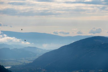 A panoramic view on endless mountain chains in Austrian Alps. The mountains are shrouded in fog. A small village among vast pastures at the bottom of the valley. Golden hour. Serenity and calmness