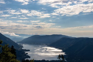 A view on the Lake Ossiacher located in between high Alpine peaks in Austria. The surface if the lake reflects golden sunbeams of the sun. A bit of overcast. Few tree brunches entering the frame