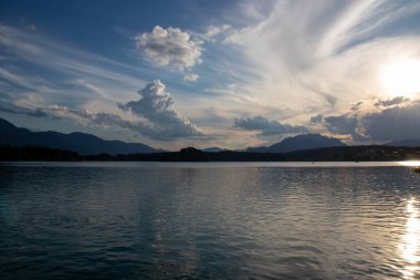 A panoramic view on the Lake Faak in Austria. The lake is surrounded by high Alpine peaks. The sun in slowly setting behind the mountains. Lots of clouds. Calm surface reflects the sunbeams. Serenity