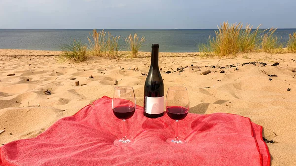 A bottle of red wine and two glasses placed on a red towel, on a sandy beach by Baltic Sea on Sobieszewo island, Poland. The beach is scarcely overgrown with high grass. A bit of overcast. Celebration
