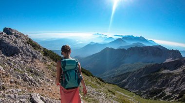 A woman with big backpack hiking along a steep pathway to the metal cross with a panoramic view on the Alps on top of Mittagskogel in Austria. Sunny day. A sun glow above the valley. Outdoor activity