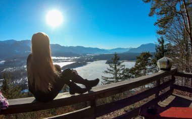 A woman in black sitting at the railing of a viewing platform and flipping her long, brown hair. There is a frozen Faaker lake underneath, surrounded by high Austrian Alps. The woman enjoys sunny day