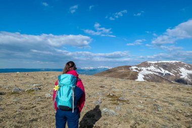 A woman hiking to the top of Ameringkogel in Austrian Alps. There is still some snow on the slopes. The vast pasture is golden. There are other chains in the back. A narrow pathway leading to the top