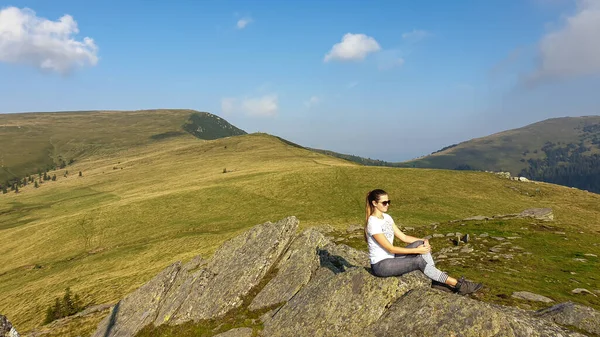 Woman in hiking outfit sitting at a big rock and enjoys the view, while hiking to Speikkogel, Austrian Alps. The mountains around are lush green, not so sharp. Sunny summer day. Serenity and calmness