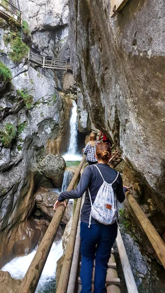 A woman in black blouse walking along a wooden, ladder pathway along a high gorge in Austrian Alps. There is a waterfall in front of her. Adventure park. Blue sky above.