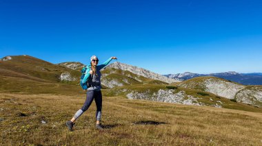 A woman with a hiking backpack hiking pointing with her finger to the peak of Hohe Weichsel in Austria. The peak is her destination. Lush green pasture around her. Exploration and discovery
