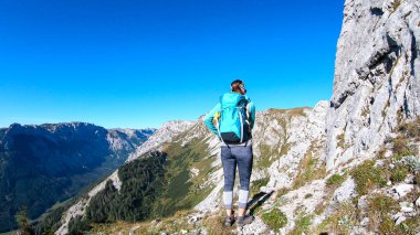 A woman with a hiking backpack climbing to the of a big boulder on the way to Hohe Weichsel in Austria, with a panoramic view on a vast valley.  Narrow pathway. She is enjoying the view. Discovering