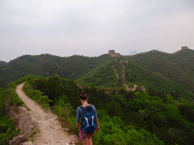 A woman with hiking backpack walking around a unrenewed Gubeikou part of Great Wall of China. The wall is spreading on tops of mountains. Dense forest around it. World wonder. Tradition and history