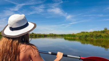 A woman in a white hat kayaking on Nida river in Poland. She is holding a red paddle in her hands. The banks of the river are overgrown with lush green grass. Clear, blue sky. Outdoor activity
