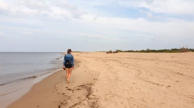 A woman with braided hair walking along the coastal line of a sandy beach by Baltic Sea on Sobieszewo island, Poland. The sea is gently waving. A bit of overcast. Solitude and serenity