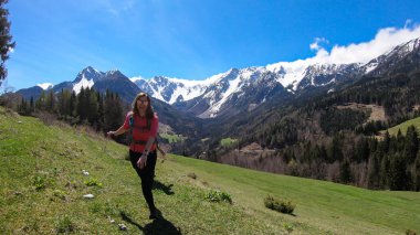 A woman with a big hiking backpack dancing with the panoramic view on Baeren Valley in Austrian Alps. The highest peaks are snow-capped. Lush green pasture. Clear and sunny day. High mountain chains