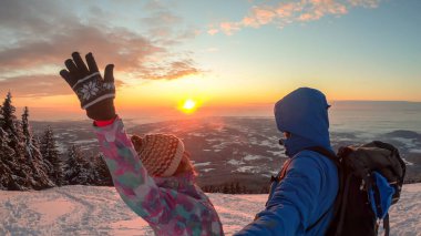 A couple enjoying the sunrise seen from snow-capped peak of Schoeckl in Austrian Alps. The sky is bursting with orange and pink. The sun rises above the horizon. Winter wonderland. Day break. Happiness