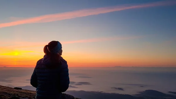 A woman with a headband sitting on top of Babia Gora, Poland, and enjoying the panoramic view on sun rising above the horizon. There are thick clouds below. The sky is pink and orange. Calmness