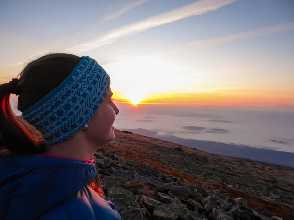A woman with a headband sitting on top of Babia Gora, Poland, and enjoying the panoramic view on sun rising above the horizon. There are thick clouds below. The sky is pink and orange. Calmness