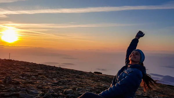 A woman with a headband dancing on top of Babia Gora, Poland, and enjoying the panoramic view on sun rising above the horizon. There are thick clouds below. The sky is pink and orange. Happiness