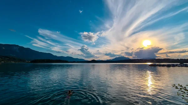 A woman swimming in the Lake Faak in Austria. The lake is surrounded by high Alpine peaks. The sun in slowly setting behind the mountains. Lots of clouds. Calm surface reflects the sunbeams. Happiness