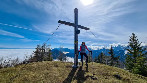 オーストリアのアルパインピークの上に十字架の横に立ってハイキング服を着た女性 周りには高いアルパインチェーンがあり 部分的に雪で覆われています 斜面に鬱蒼とした森 晴れた日だ — ストック写真