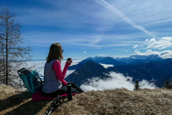A woman in hiking outfit sipping a tea from thermos with the panoramic view from the top of Alpine peak in Austria. The area is shrouded in thick clouds. A few peaks popping out from the clouds. Happy