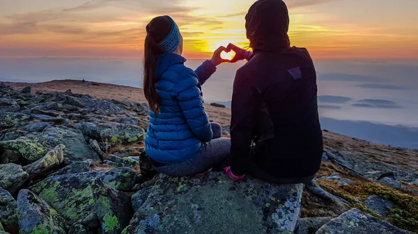 一对夫妇坐在波兰巴比亚戈拉山顶上 欣赏着地平线上升起的太阳的全景 他们用双手形成了一颗心 下面乌云密布 天空是粉红色和橙色的 — 图库照片