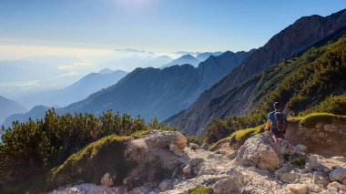 A man in a hiking outfit with a panoramic view on the haze shrouded valley from the way to Mittagskogel in Austrian Alps. Clear and sunny day. Endless mountain chains. Outdoor activity. Achievement