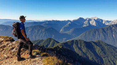 A man in a hiking outfit with a panoramic view on the haze shrouded valley from the way to Mittagskogel in Austrian Alps. Clear and sunny day. Endless mountain chains. Outdoor activity. Achievement