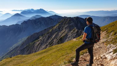 A man in a hiking outfit with a panoramic view on the haze shrouded valley from the way to Mittagskogel in Austrian Alps. Clear and sunny day. Endless mountain chains. Outdoor activity. Achievement